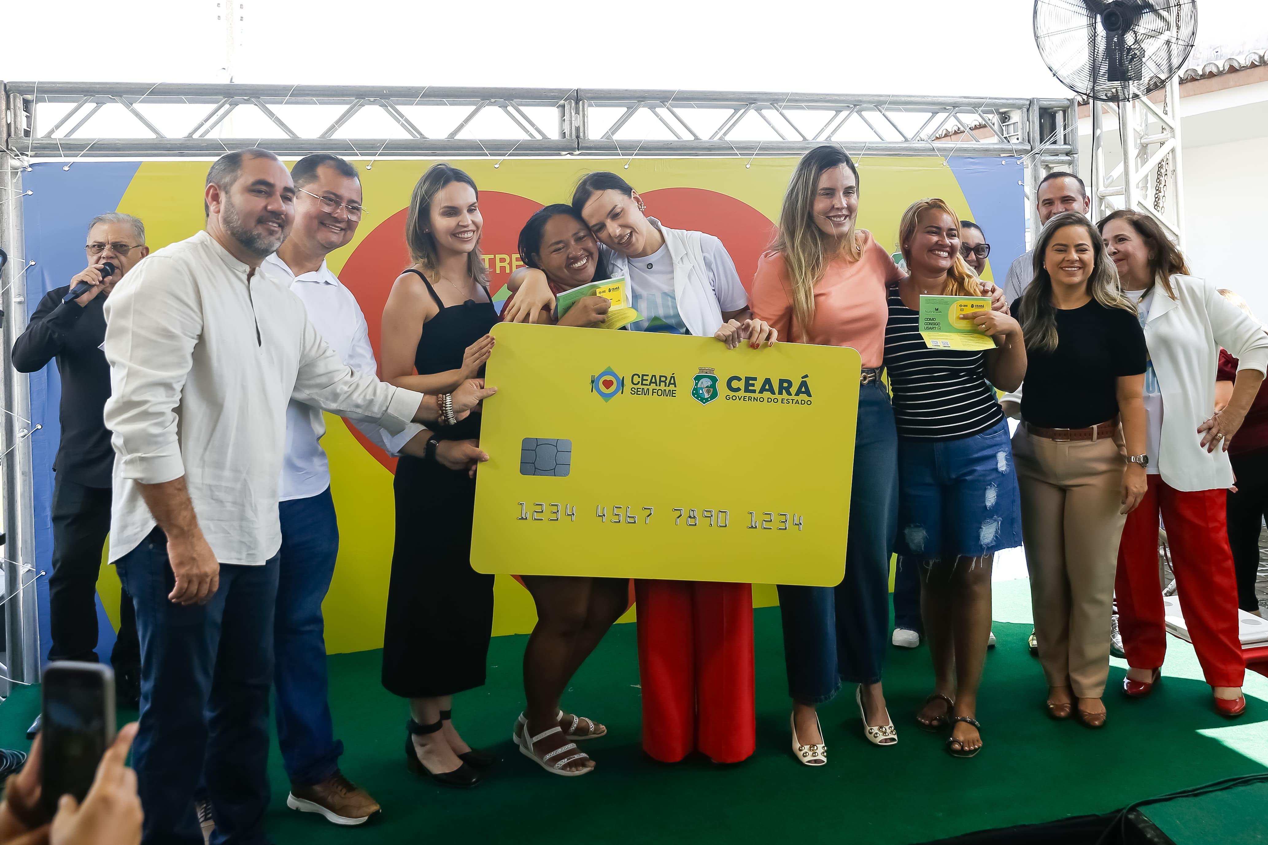 primeira-dama do estado entrega uma réplica gigante de um cartão de crédio que representa o cartão do programa ceará sem fome para umaa mulher. elas estão em um palco cercadas por outras pessoas